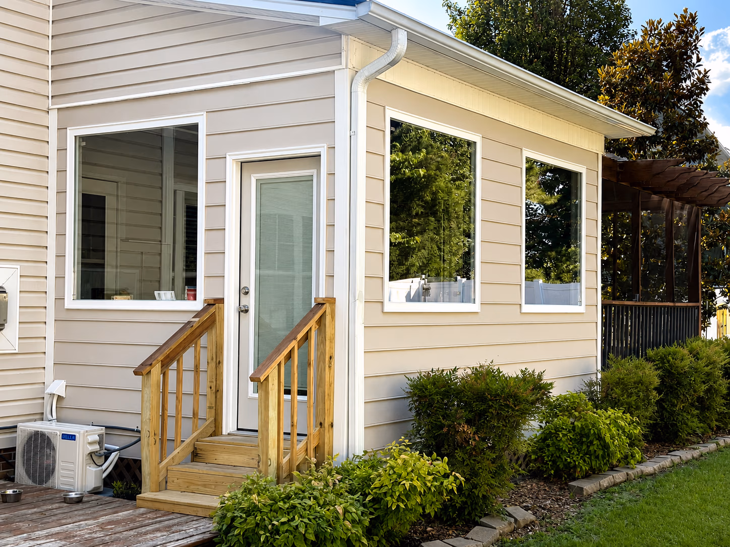Backyard view of beige house with three large windows, white door, wooden stairs, and green bushes.