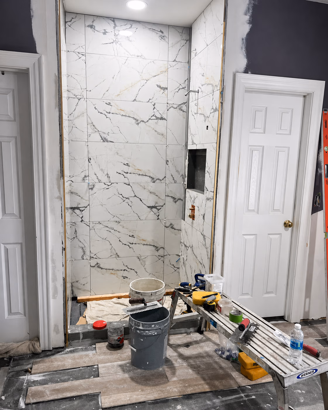 Bathroom under renovation with marble shower tiles, wood plank flooring, tools, and supplies scattered around.