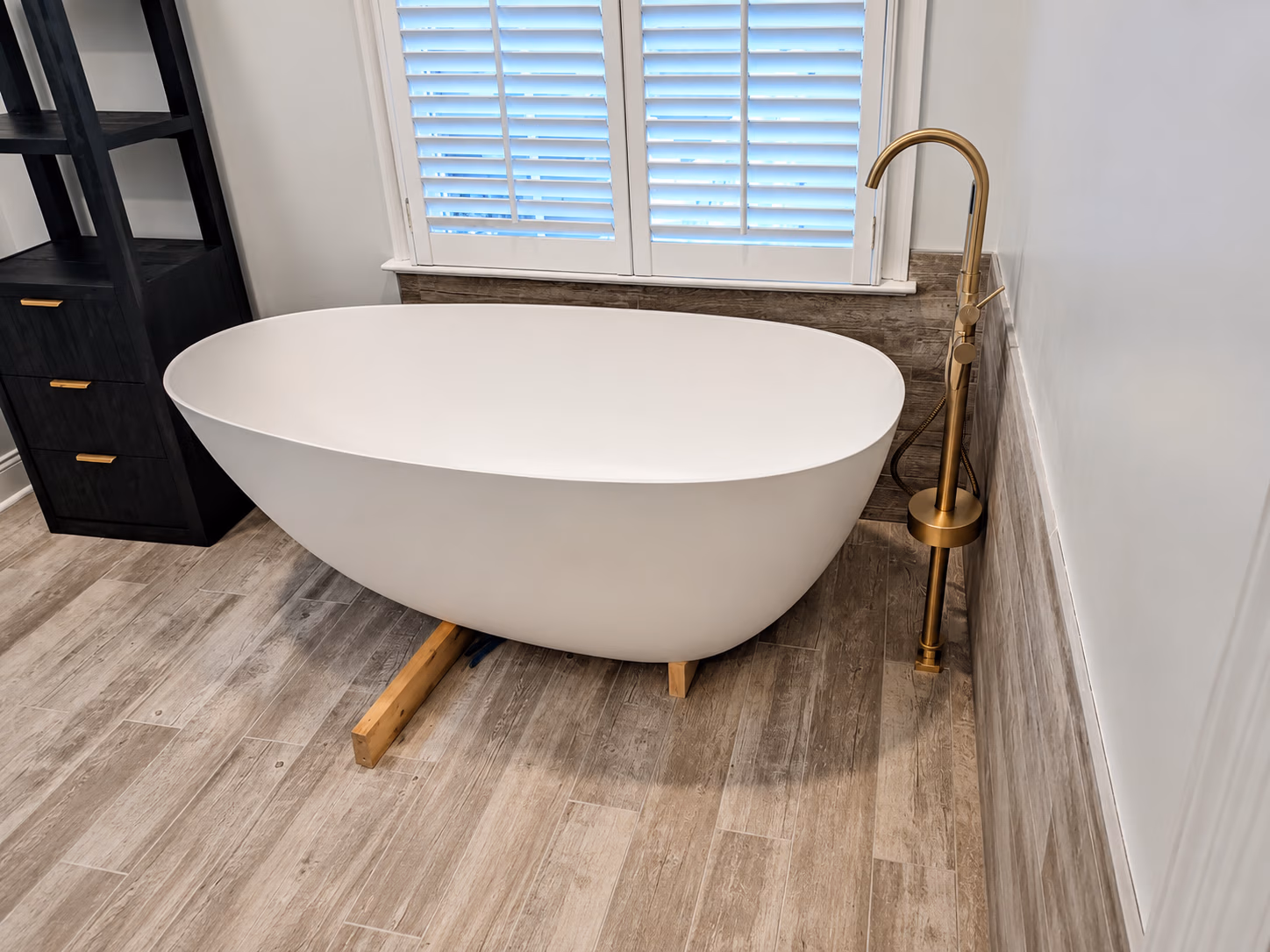 Modern white freestanding bathtub with gold faucet on wood-look tile floor by a window with shutters.
