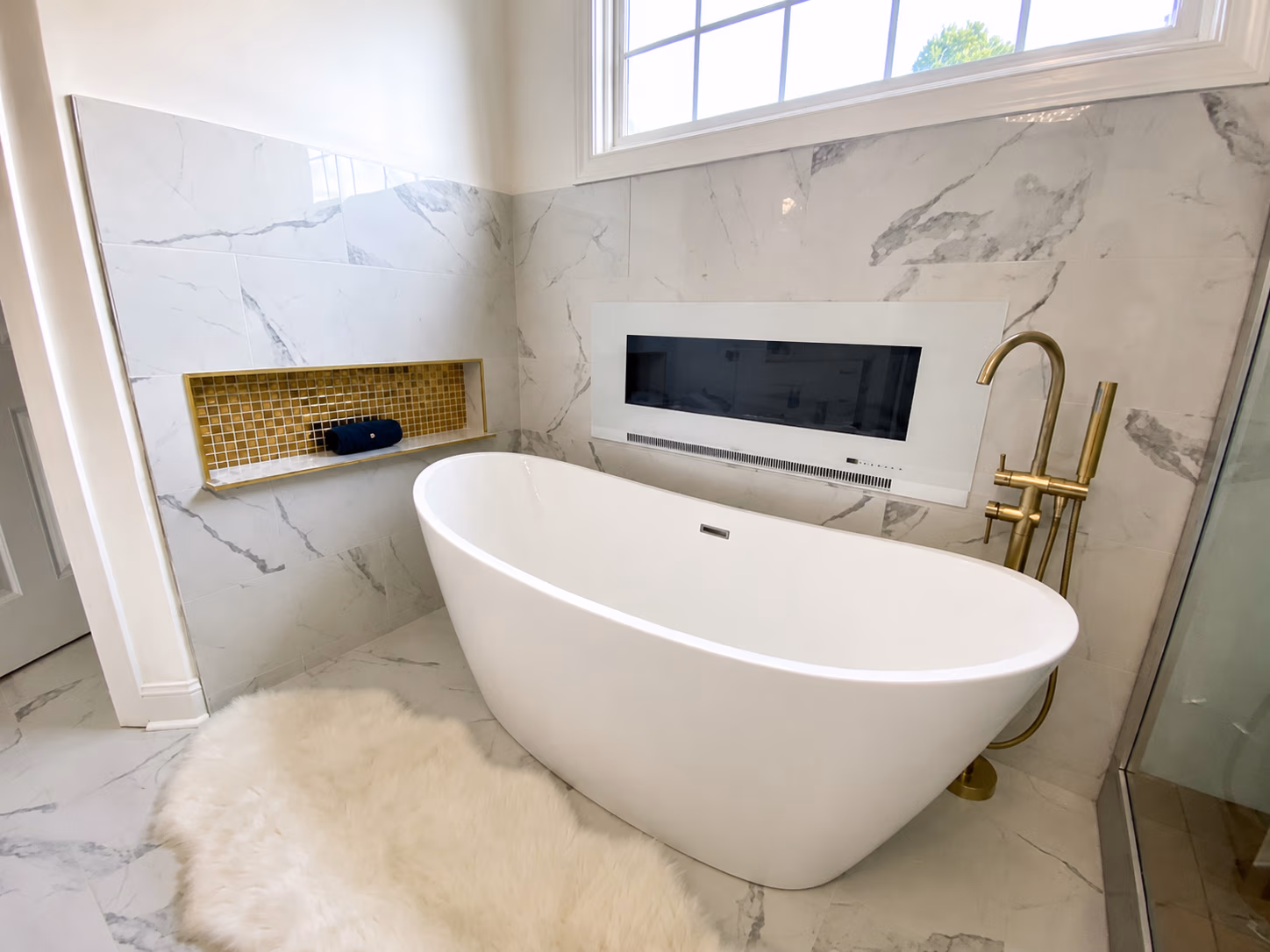 Modern bathroom with a white freestanding tub, gold faucet, and built-in electric fireplace above marble tiles.