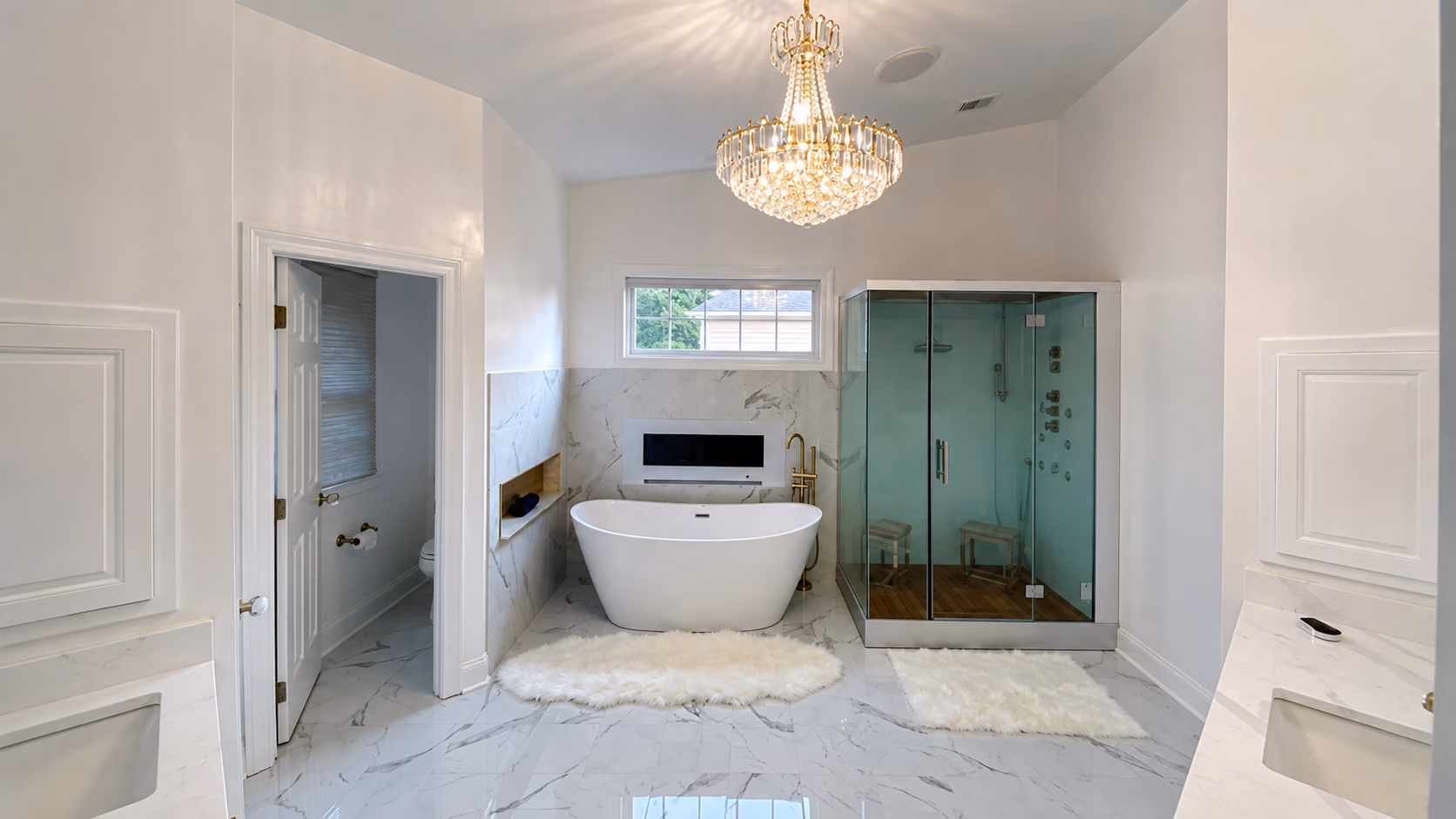Modern bathroom with freestanding bathtub, glass shower, chandelier, and marble floors with rugs.