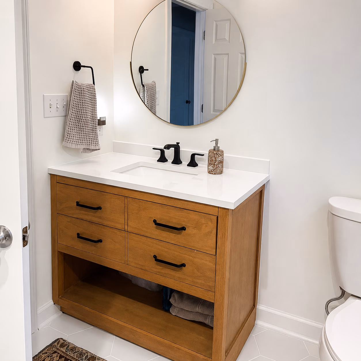 Modern bathroom with wooden vanity, white countertop, round mirror, black fixtures, and a toilet on the right.