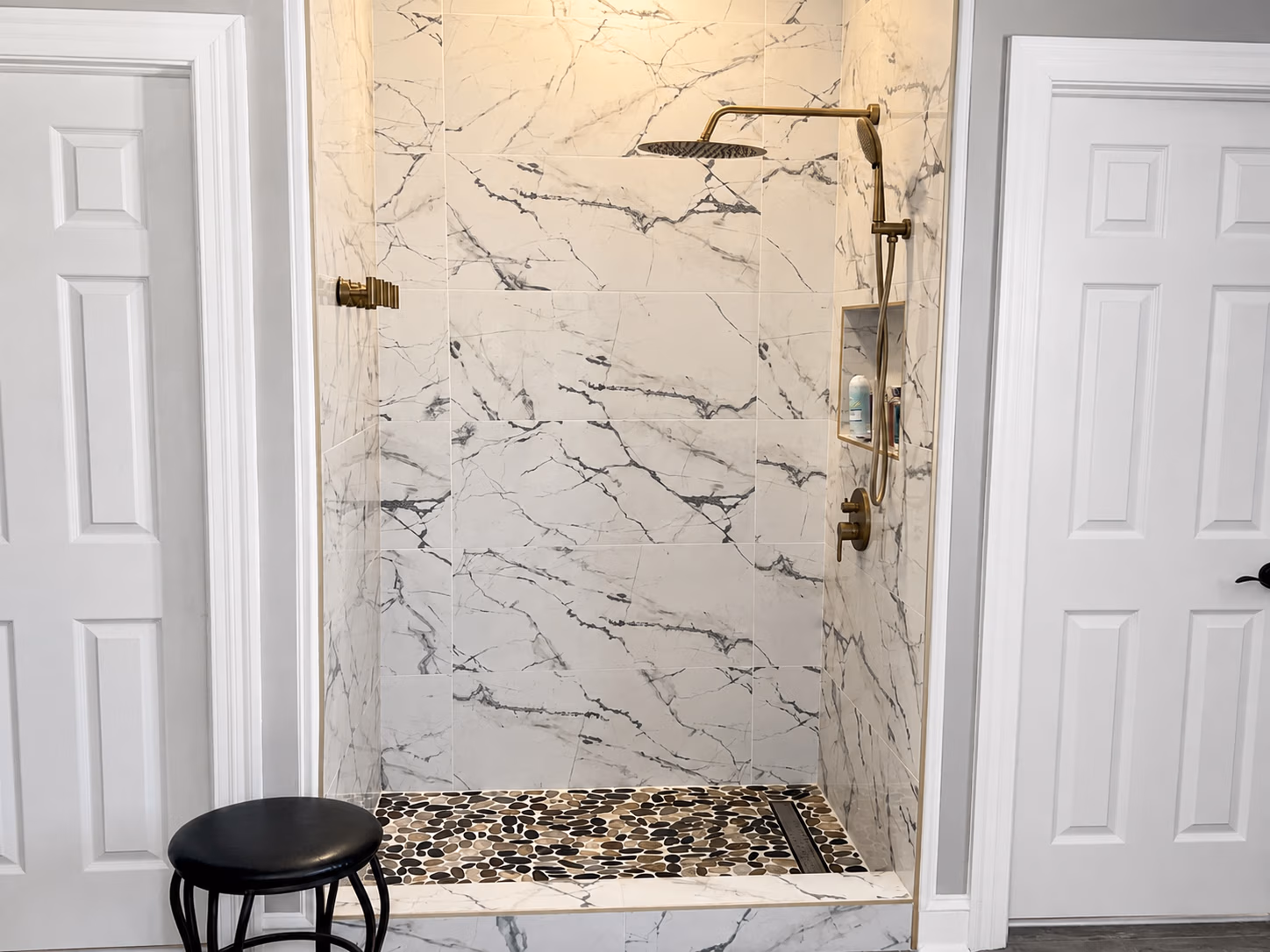 Modern walk-in shower with white marble tiles, brass fixtures, and pebble stone floor beside a black stool.