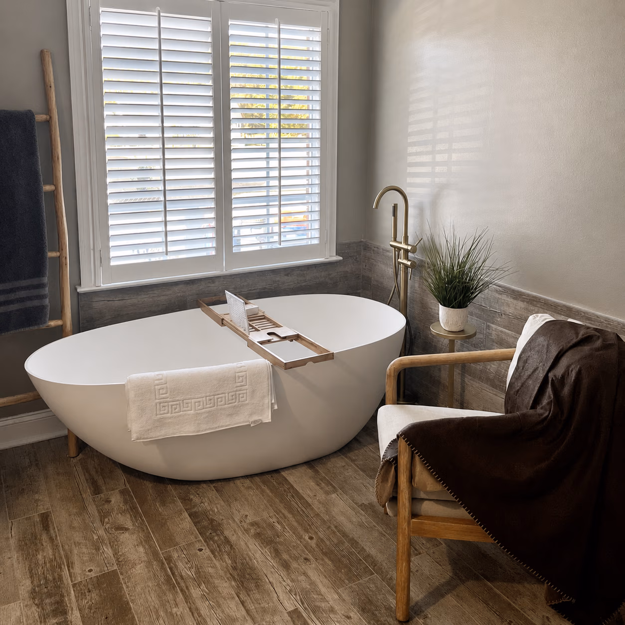 Modern bathroom with white freestanding tub, wooden floor, plant, and chair with brown blanket.