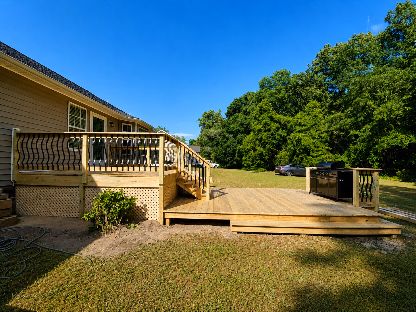 Wooden backyard deck attached to a house with black railing and an outdoor grill on a sunny day.