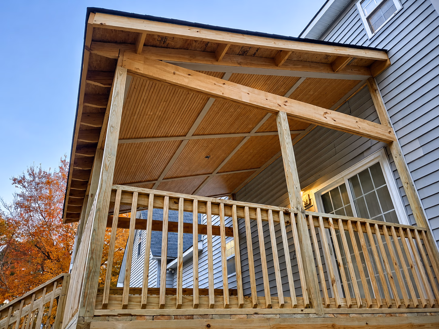 Wooden covered porch attached to a gray house with autumn trees in the background.