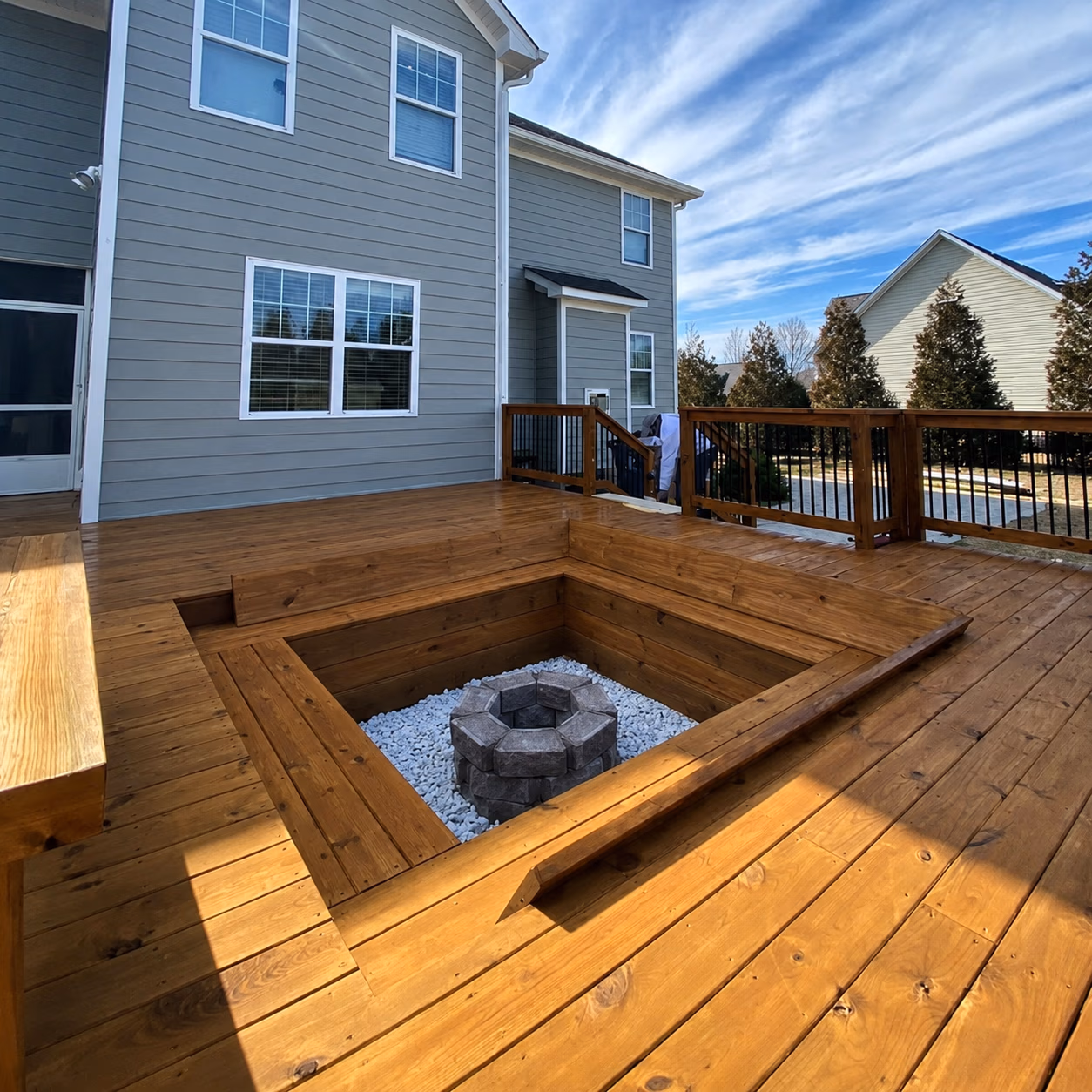Spacious wooden deck attached to a house with a built-in sunken fire pit surrounded by white stones.