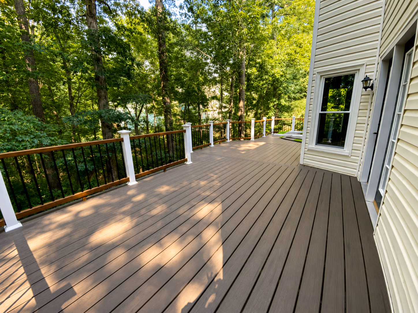 Spacious wooden deck attached to a house, surrounded by green trees and a railing.