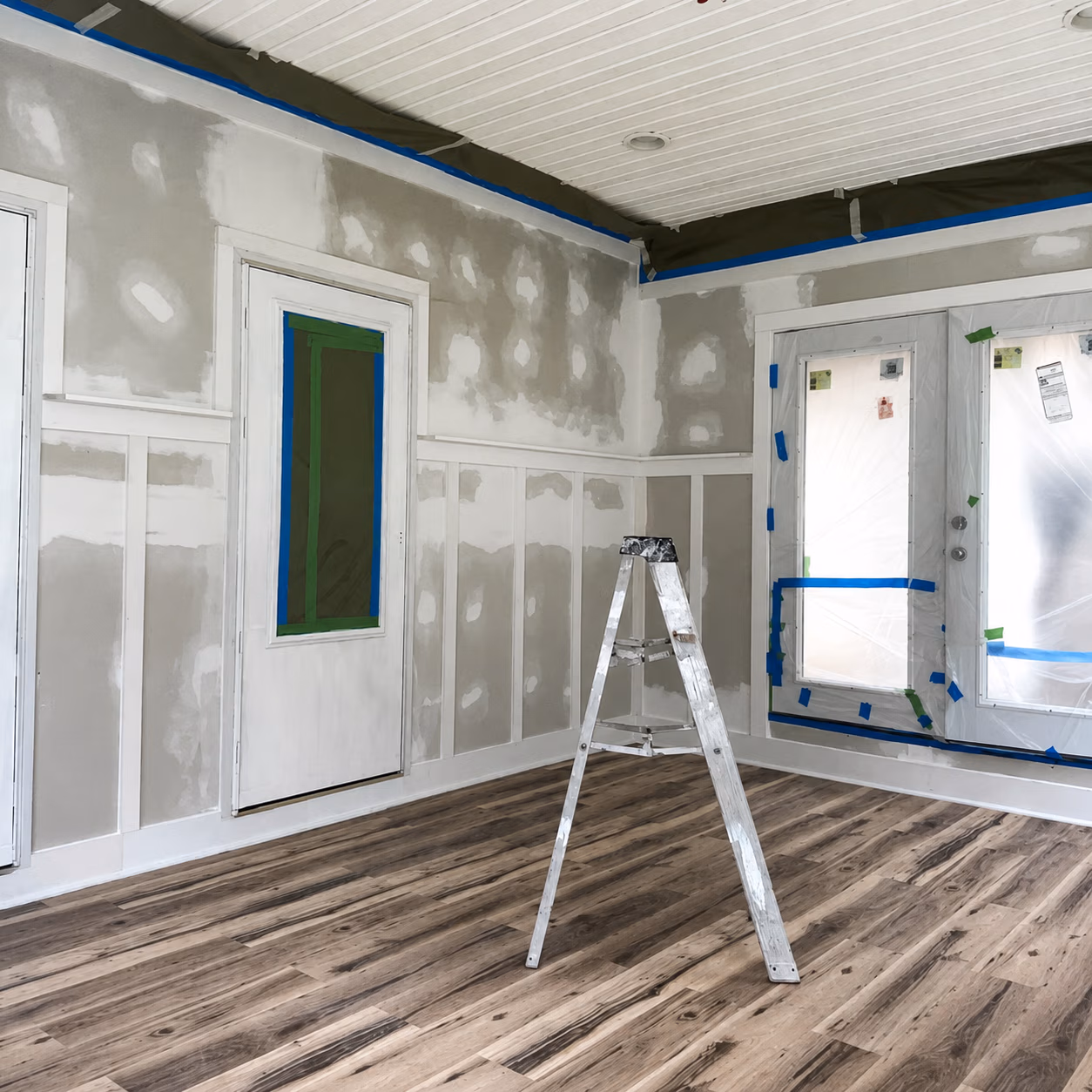 Room under renovation with patched drywall, wood floor, covered windows, and a metal step ladder.