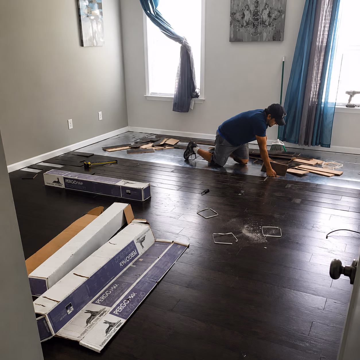 Man installing dark wood laminate flooring in a bright room with tools and boxes around him.