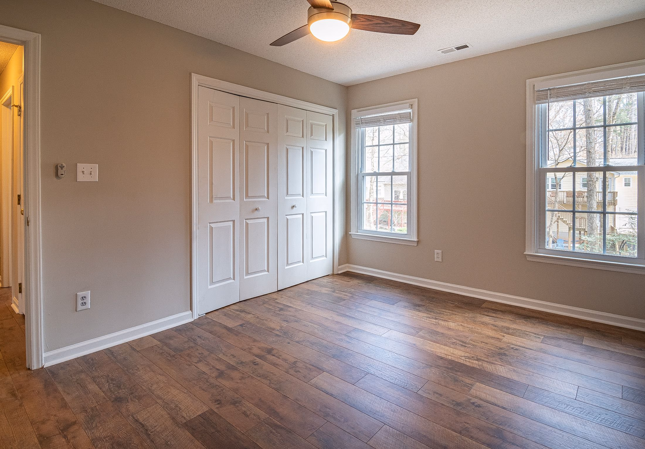 Empty room with wooden floor, white closet doors, beige walls, two windows, and ceiling fan light.