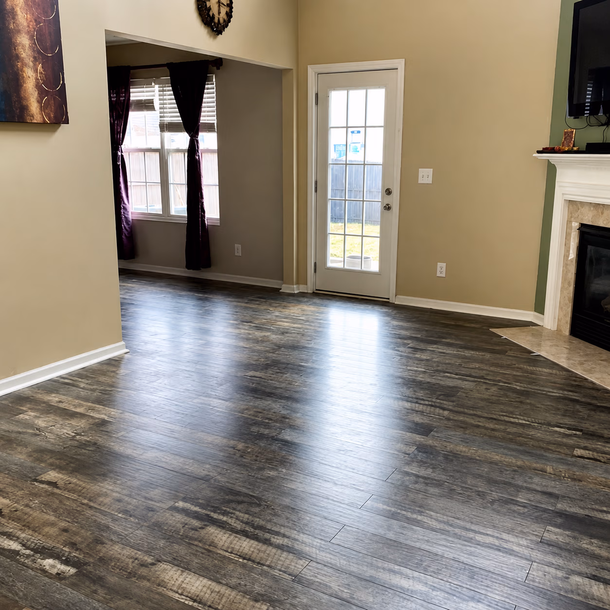 Empty room with dark wood flooring, beige walls, a glass door, window with curtains, and a fireplace.