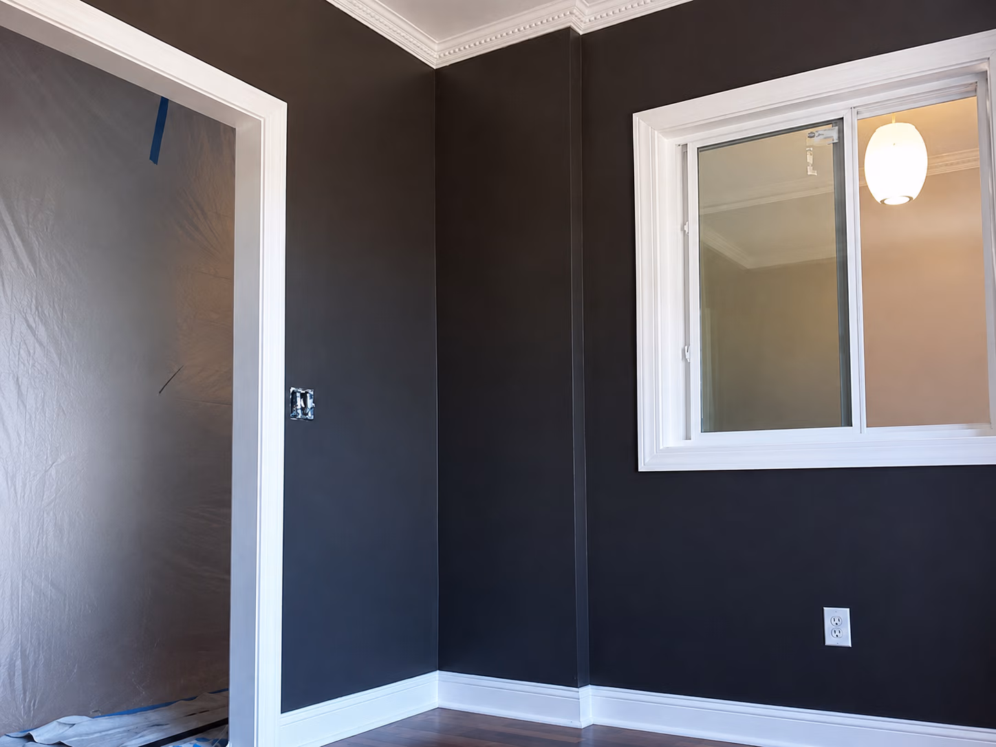 Corner of a room with dark walls, white trim, an open doorway, a window, and hardwood floor.