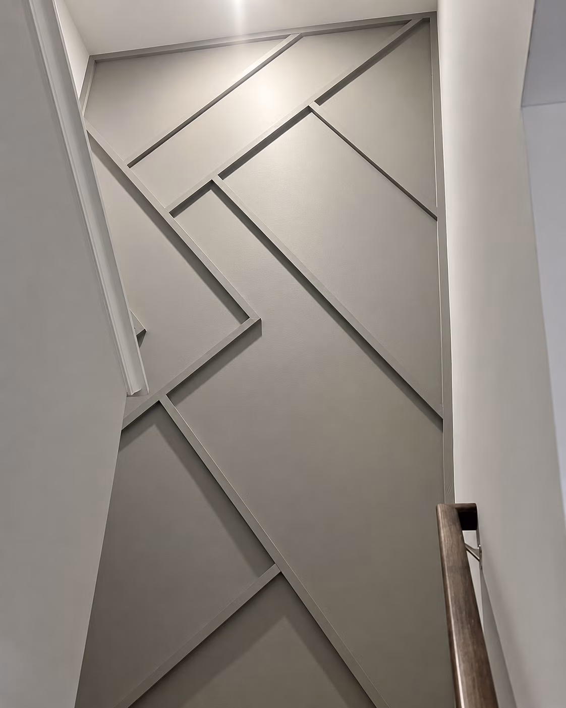 Gray wall with raised geometric paneling seen from staircase with wooden handrail.