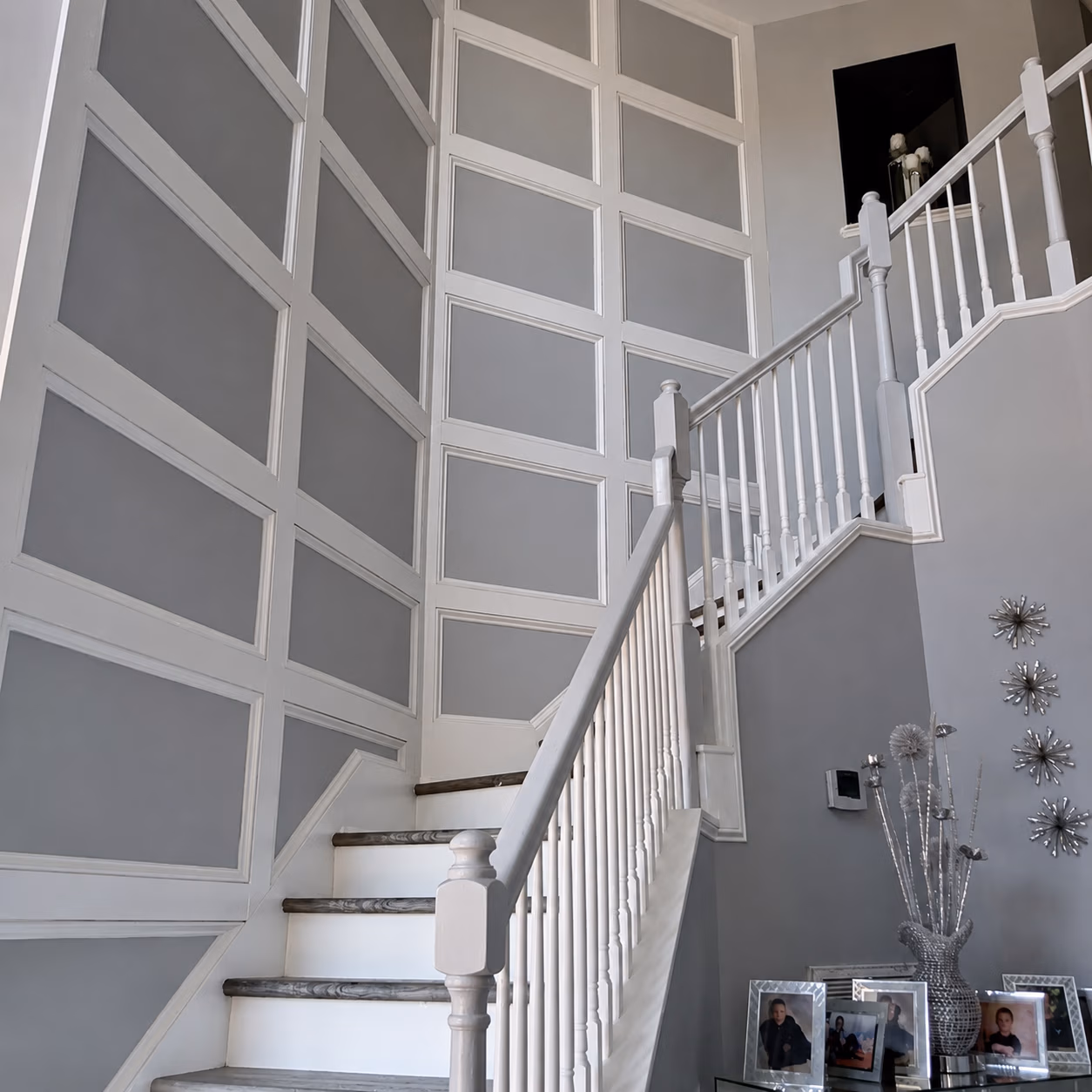 Interior staircase with white railing and gray paneled walls, beside a table with photo frames and decorative items.