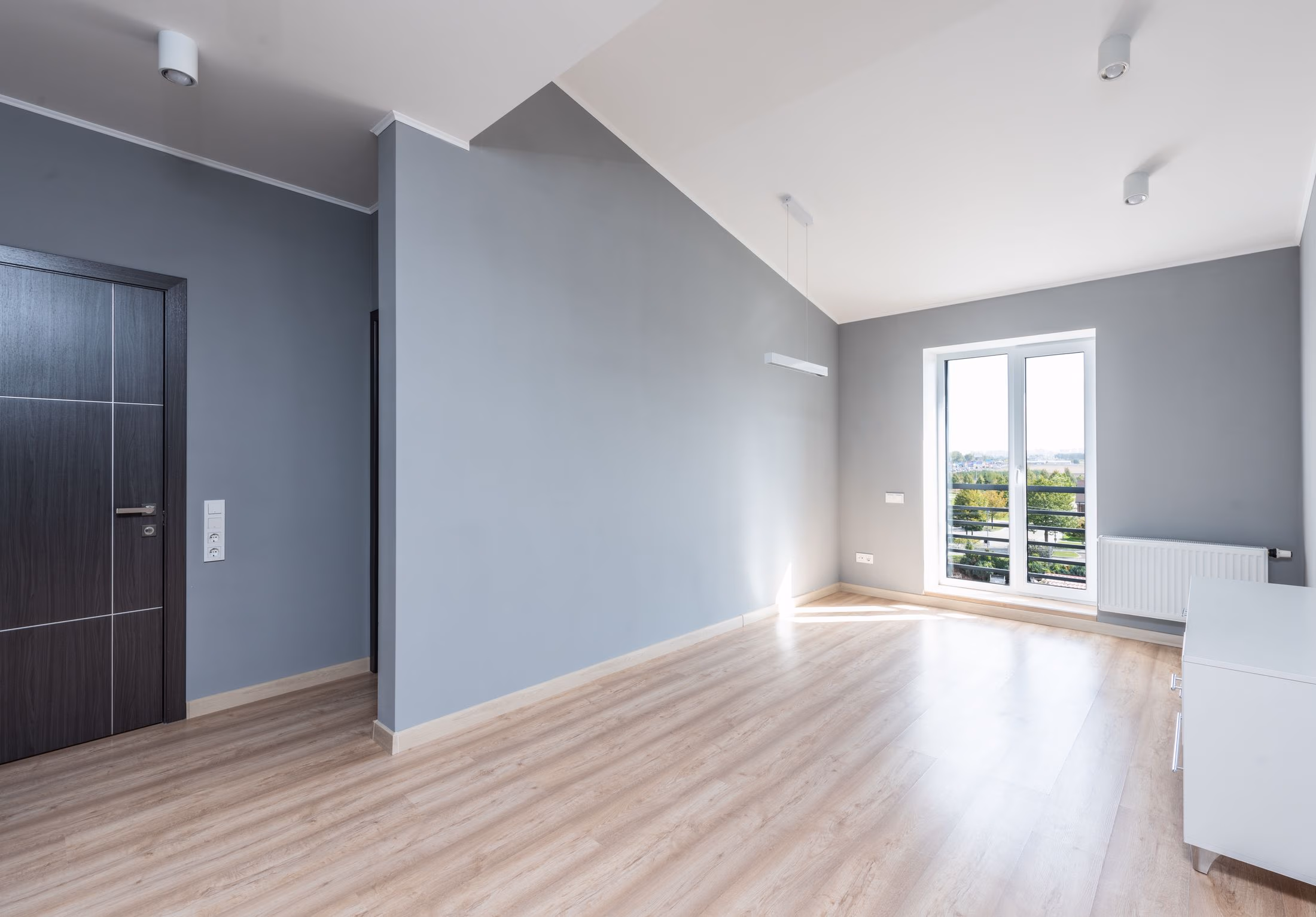 Empty modern room with gray walls, light wood floor, dark door, and large window with balcony view.