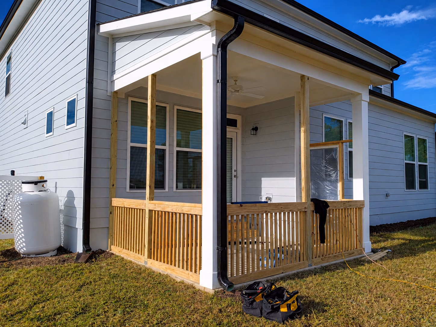 Wooden porch under construction attached to a white house with tools on grass nearby.