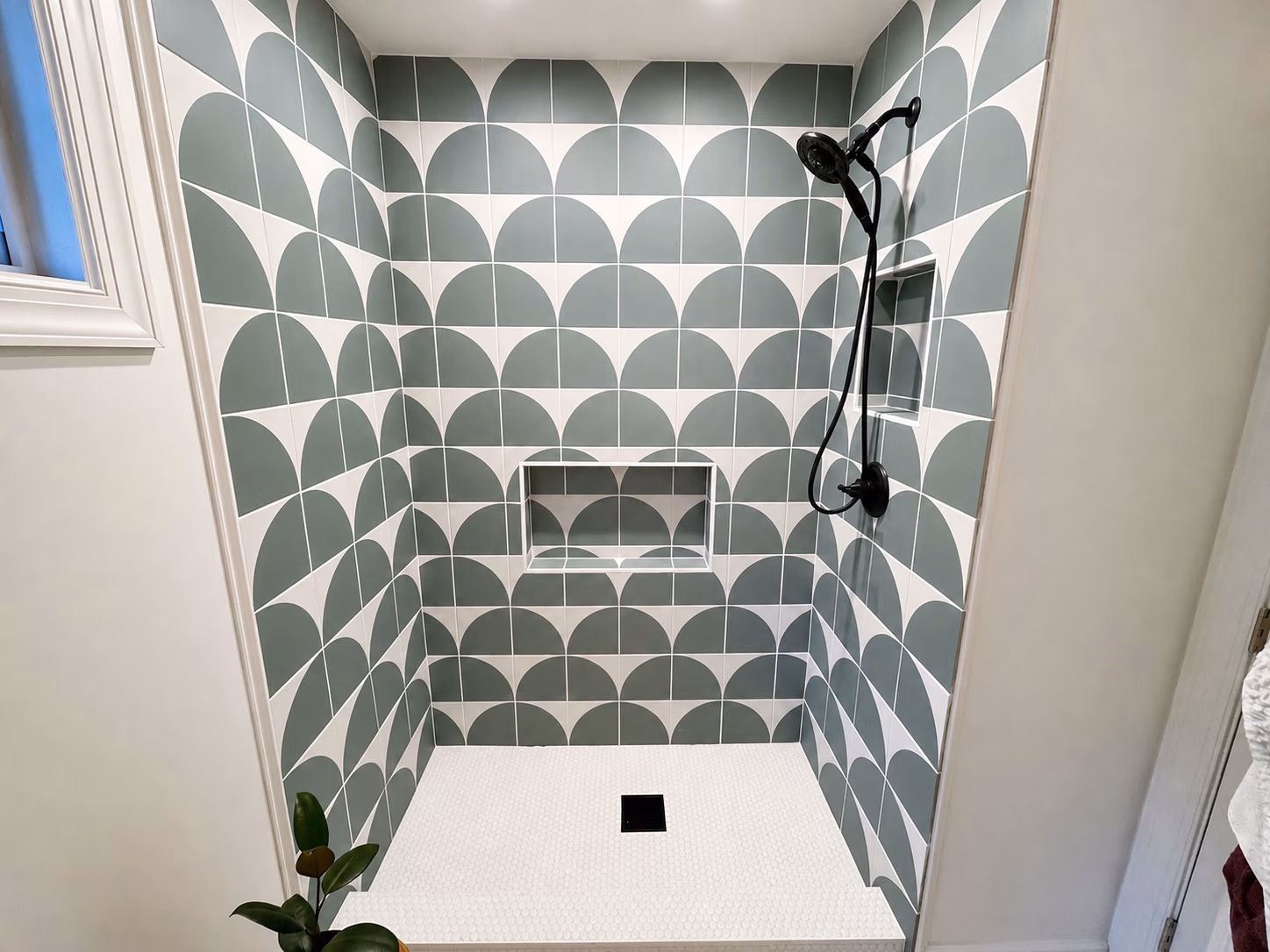 Shower with green and white geometric tiles, black showerhead, built-in shelves, and a small plant.