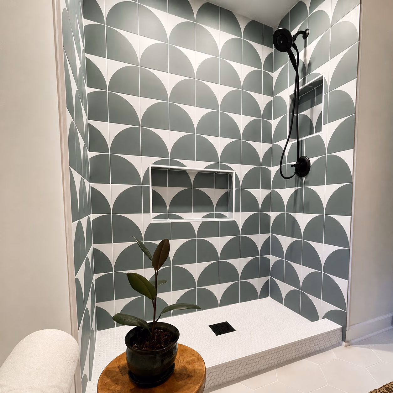 Shower with geometric green and white tiles, black showerhead, built-in shelf, and plant on wooden stool.