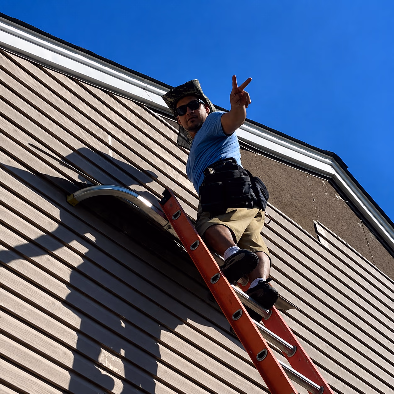 Worker in hat and sunglasses on ladder fixing tan siding on house under clear blue sky.