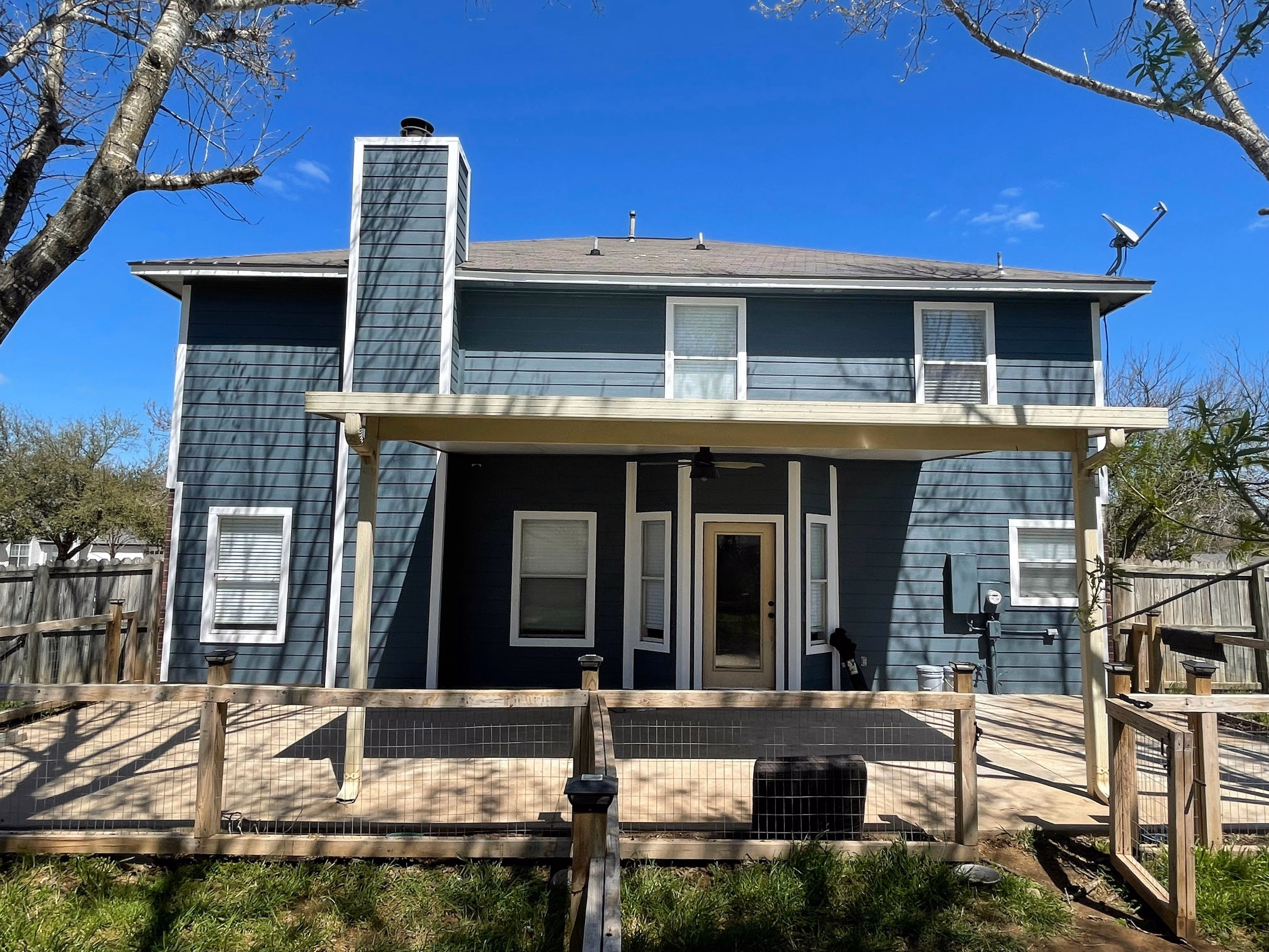 Two-story blue house with white trim, covered patio, chimney, and wooden fenced yard under clear sky.