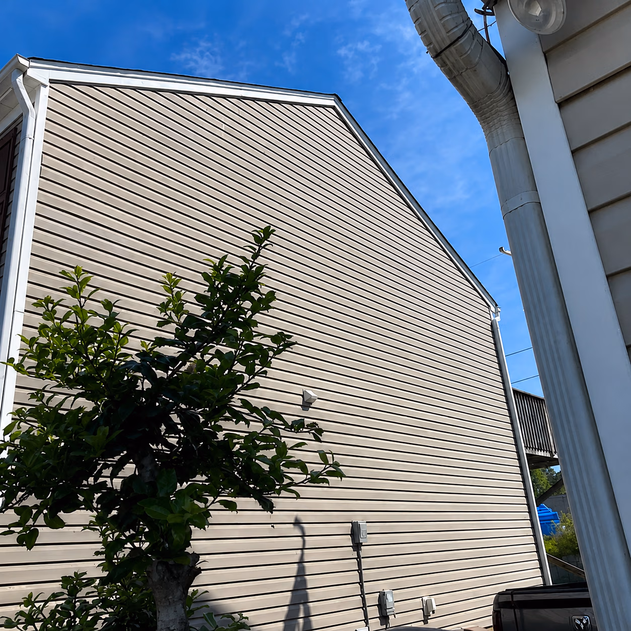 Side of a beige house with horizontal siding, a small tree, and a blue sky above.