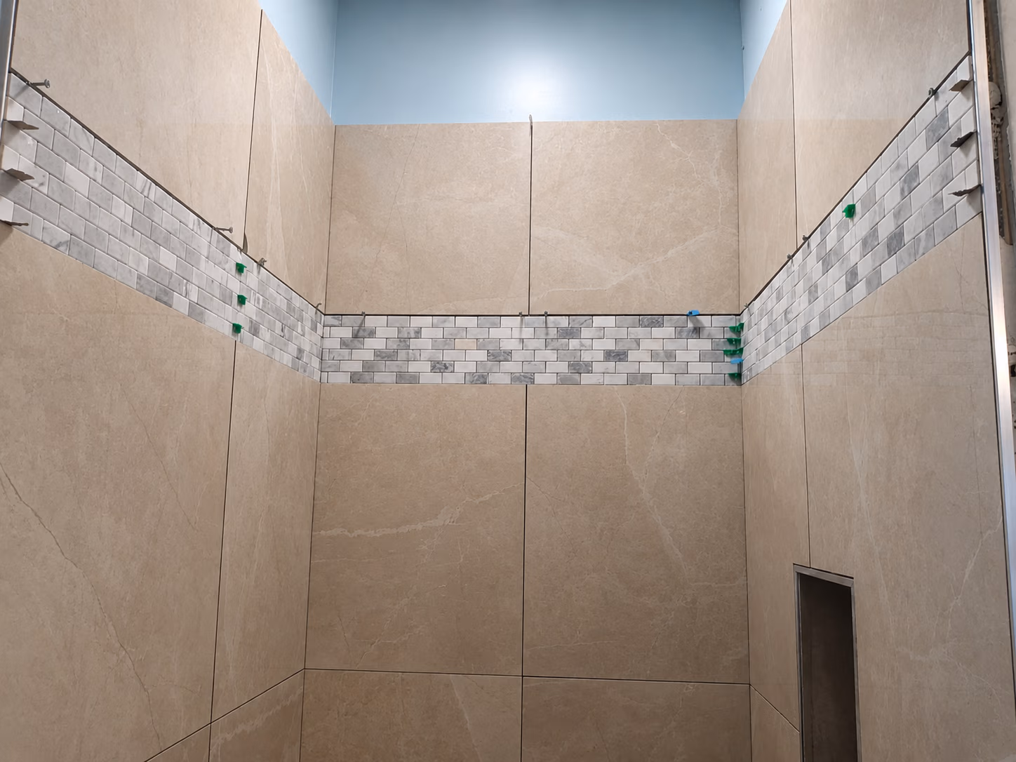 Shower walls tiled in beige with a horizontal band of small gray and white tiles and a recessed niche.