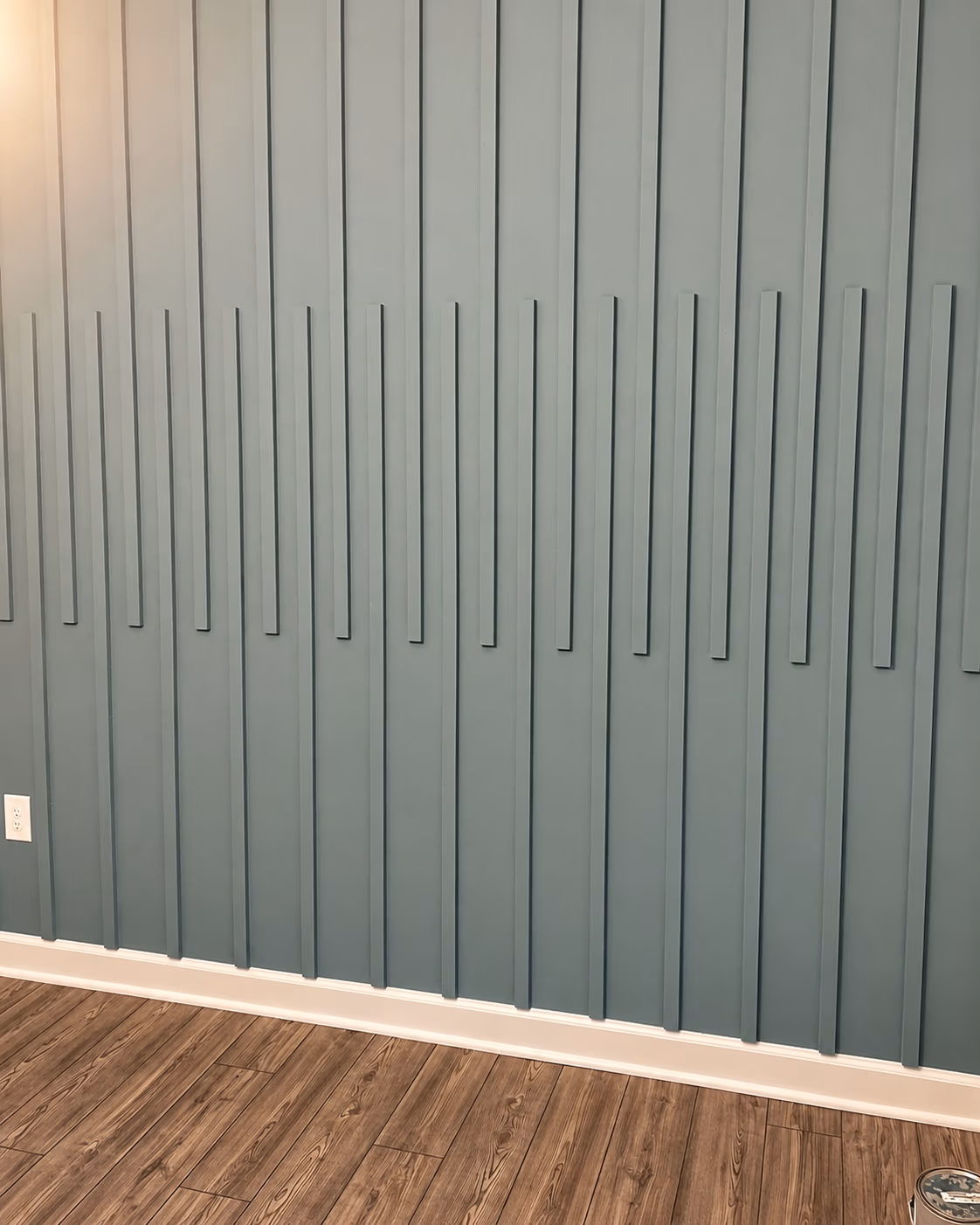 Blue wall with vertical decorative wood strips, white baseboard, brown wooden floor, and an electrical outlet.